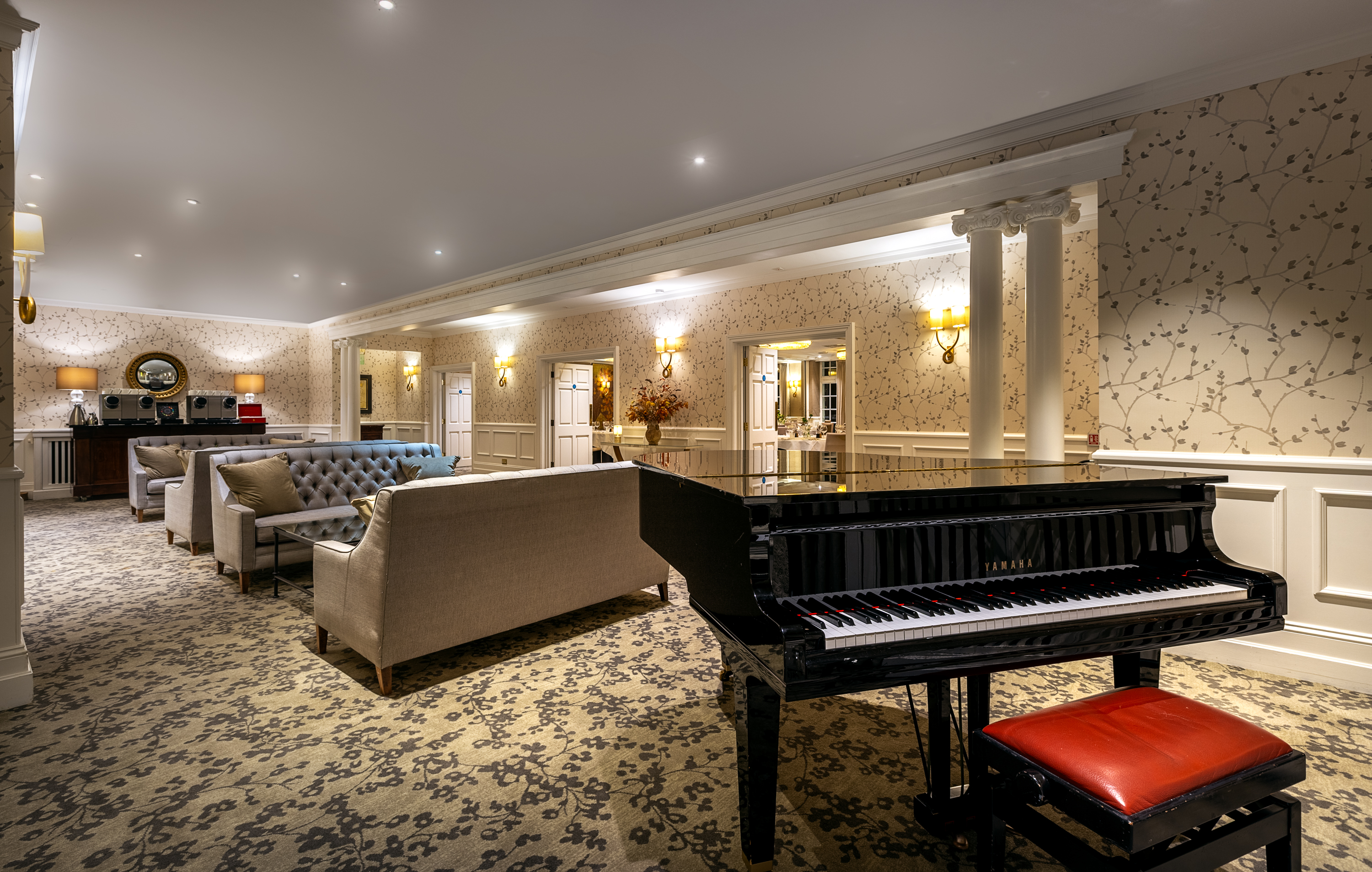 Elegant lounge with patterned walls, soft lighting, upholstered sofas, and a black grand piano with a red bench