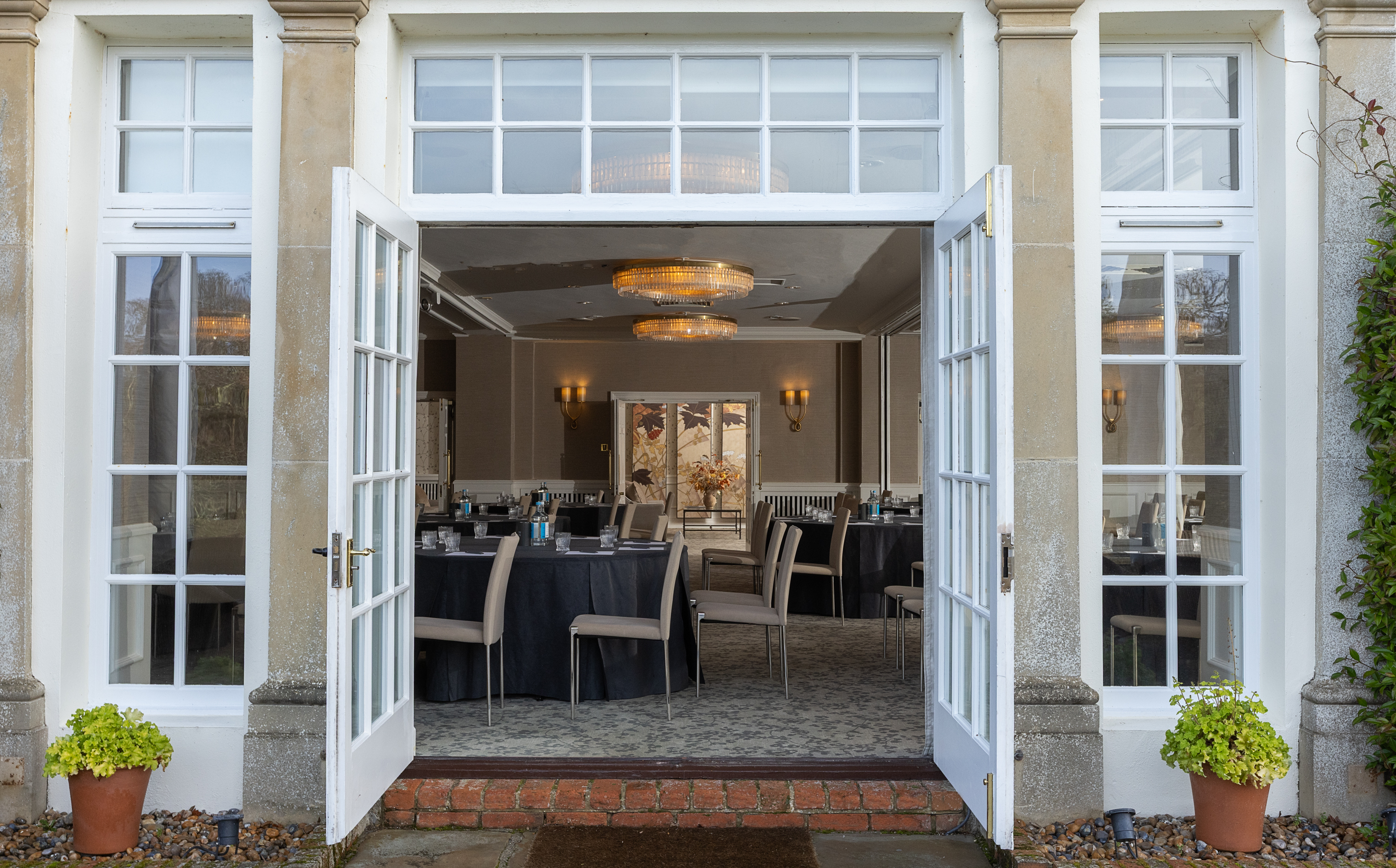 Open French doors leading into an elegant event room set with round tables, neutral‑tone chairs, and warm chandelier lighting.