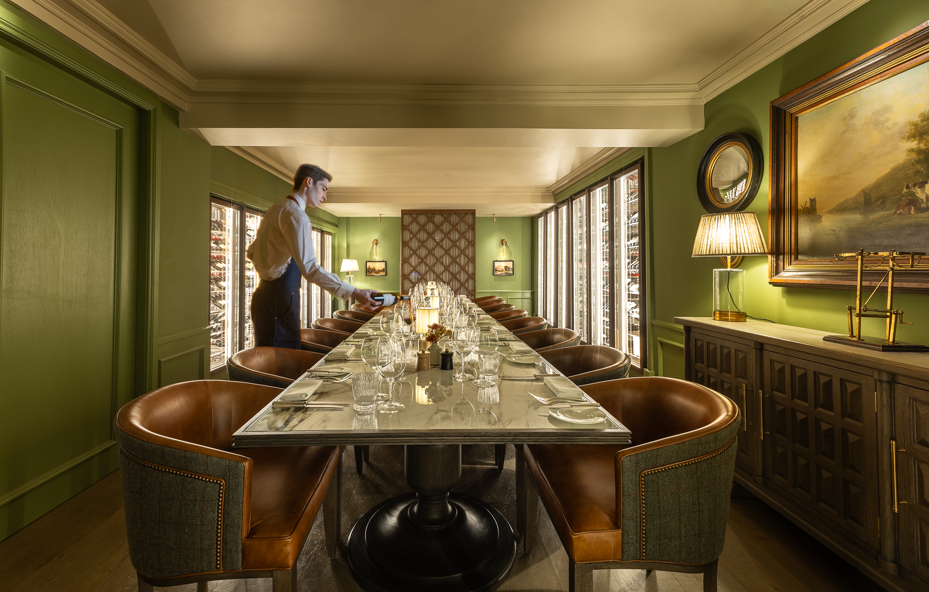 Elegant private dining room with a long marble table set with glassware and cutlery, surrounded by leather chairs, with green walls, artwork, and illuminated wine cabinets; a staff member sets the table