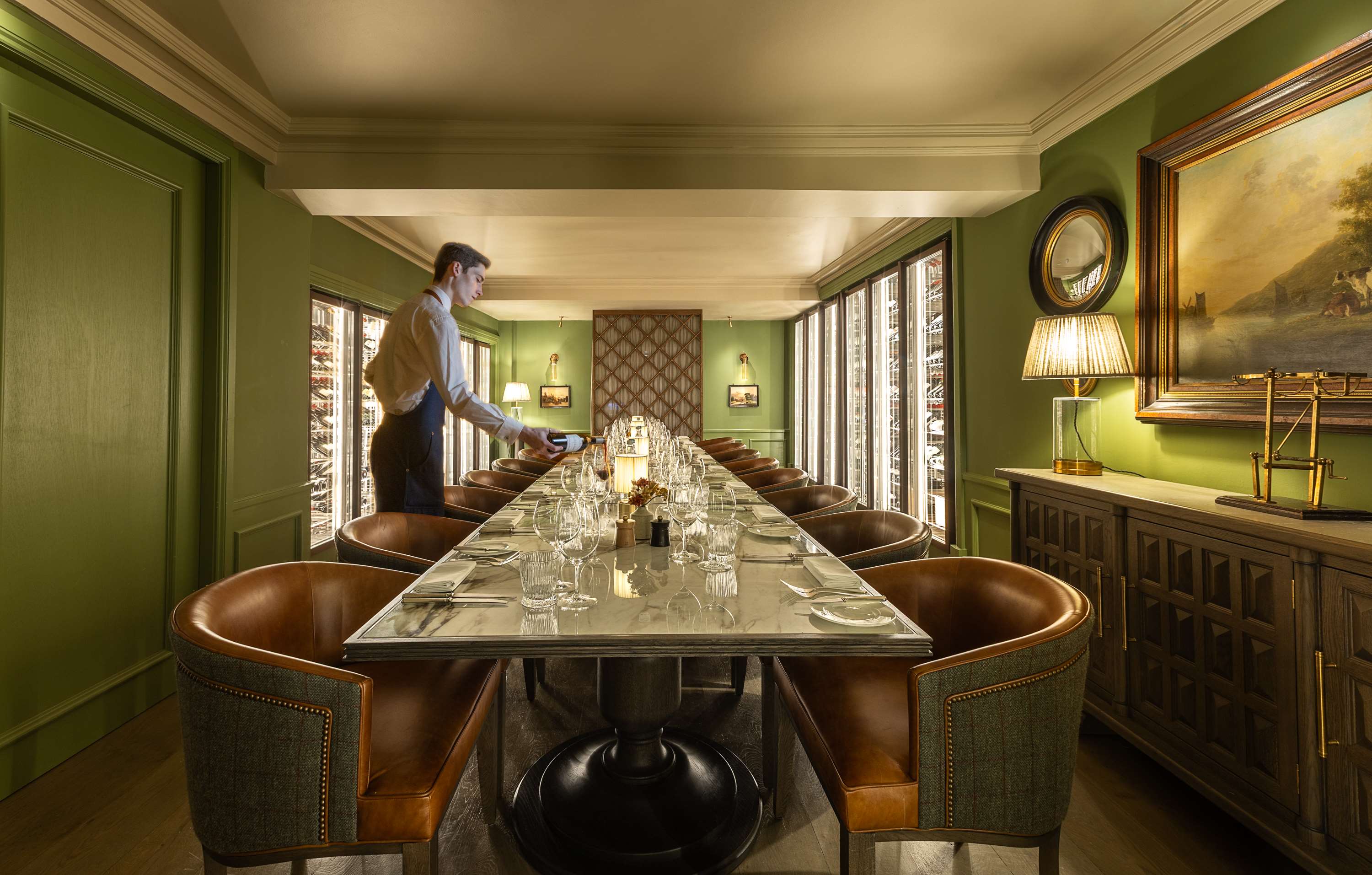 Elegant private dining room with a long marble table set with glassware and cutlery, surrounded by leather chairs, with green walls, artwork, and illuminated wine cabinets; a staff member sets the table