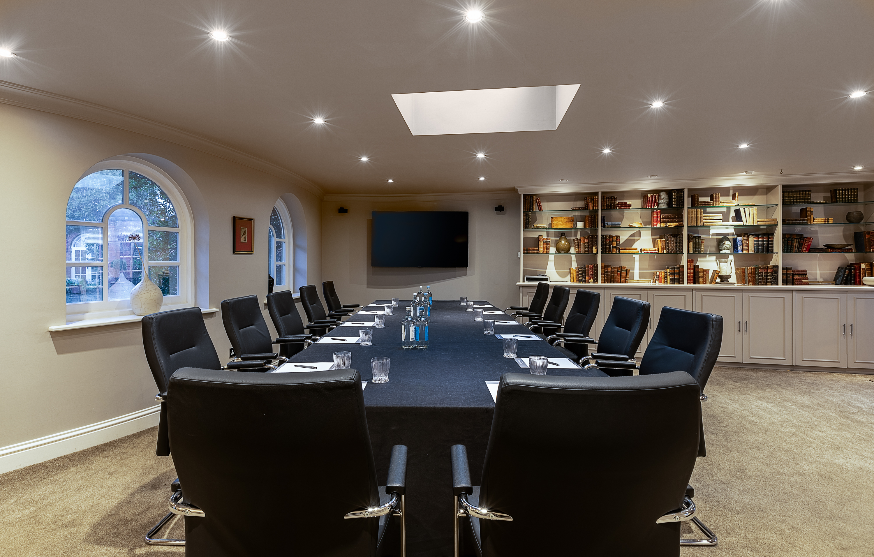 Boardroom with a long table set with notepads, glasses, and water bottles, surrounded by black chairs, with bookshelves and arched windows providing natural light