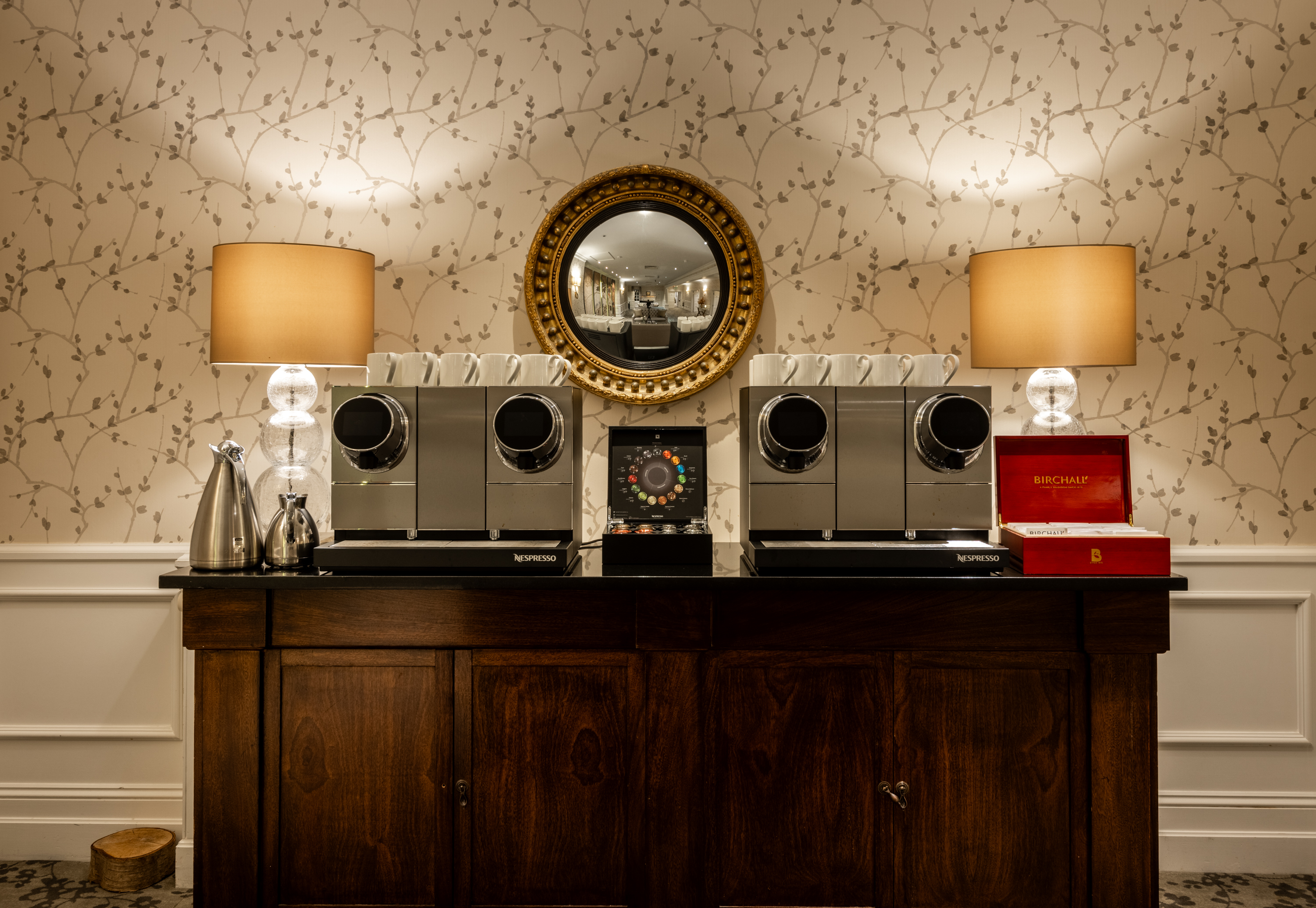 Coffee station with multiple espresso machines on a dark wooden cabinet, flanked by two lamps and a round gold-framed mirror on a patterned wall