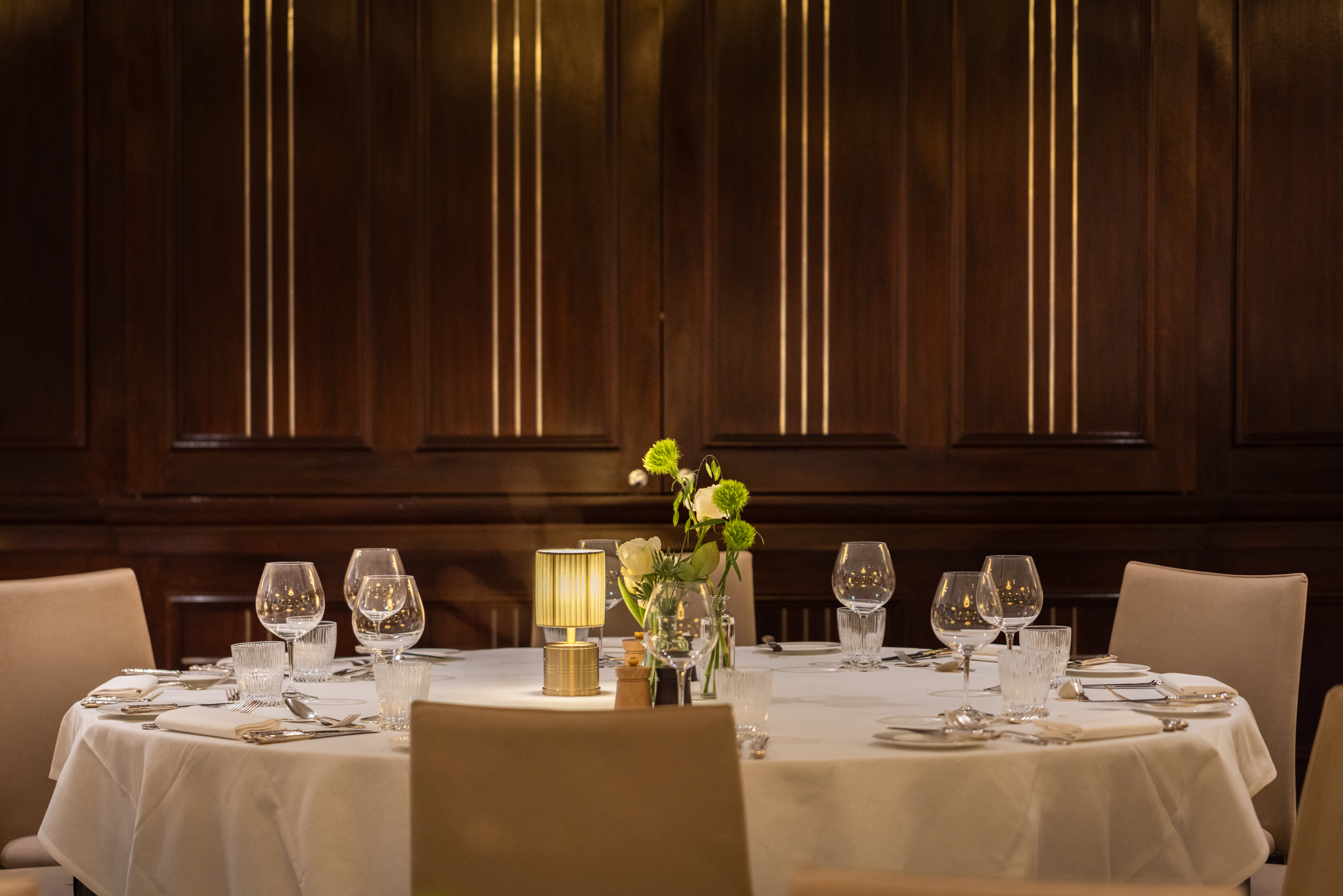 A round dining table set for a formal meal, featuring neatly arranged glassware, cutlery, a small lamp, and a floral centerpiece, with soft lighting against a dark wooden paneled wall.