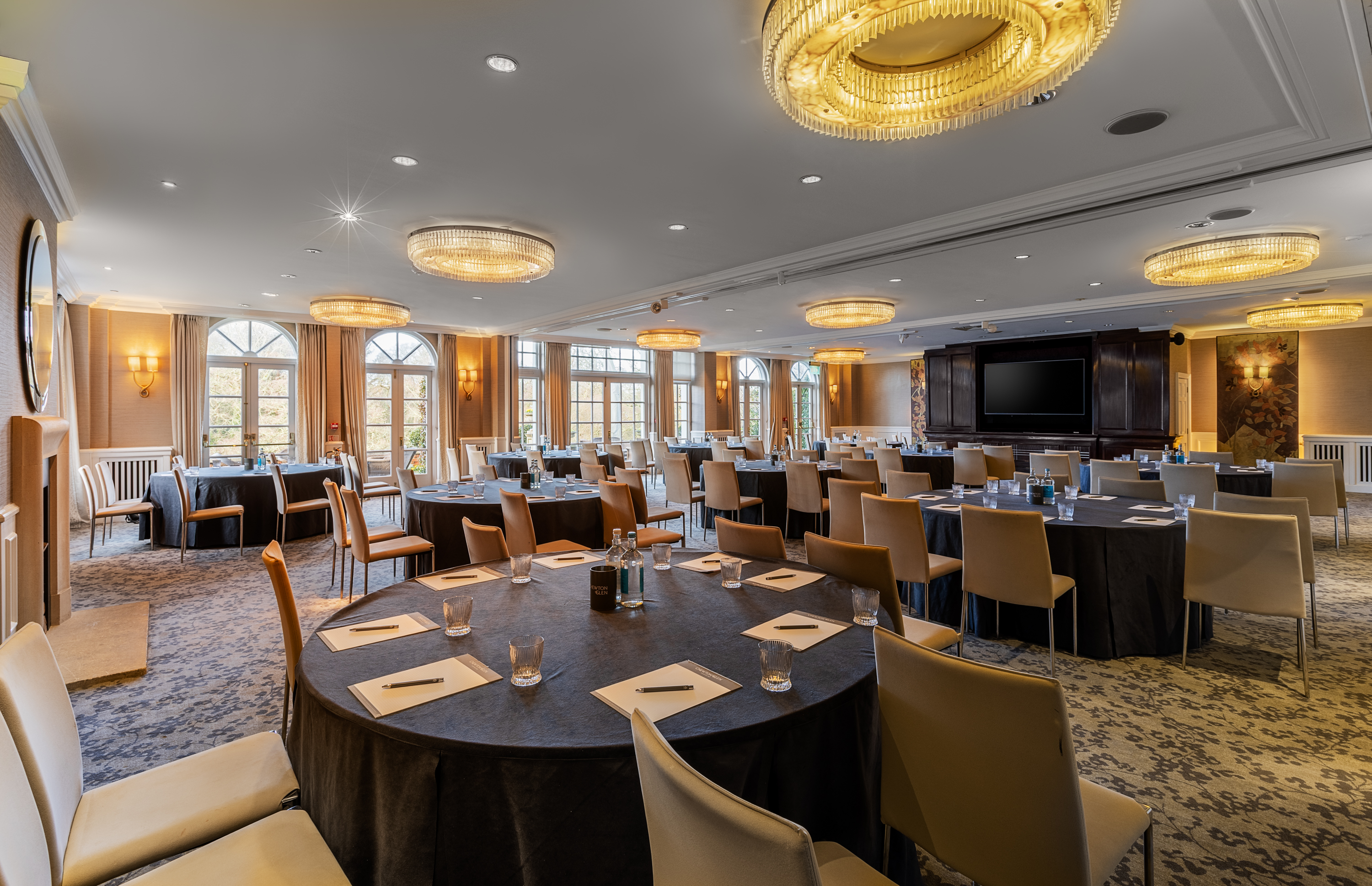 A spacious, elegant function room set up with round and rectangular tables prepared for a meeting or event, featuring notepads, glasses, and chairs throughout, with large windows, patterned carpet, and multiple chandelier-style ceiling lights.