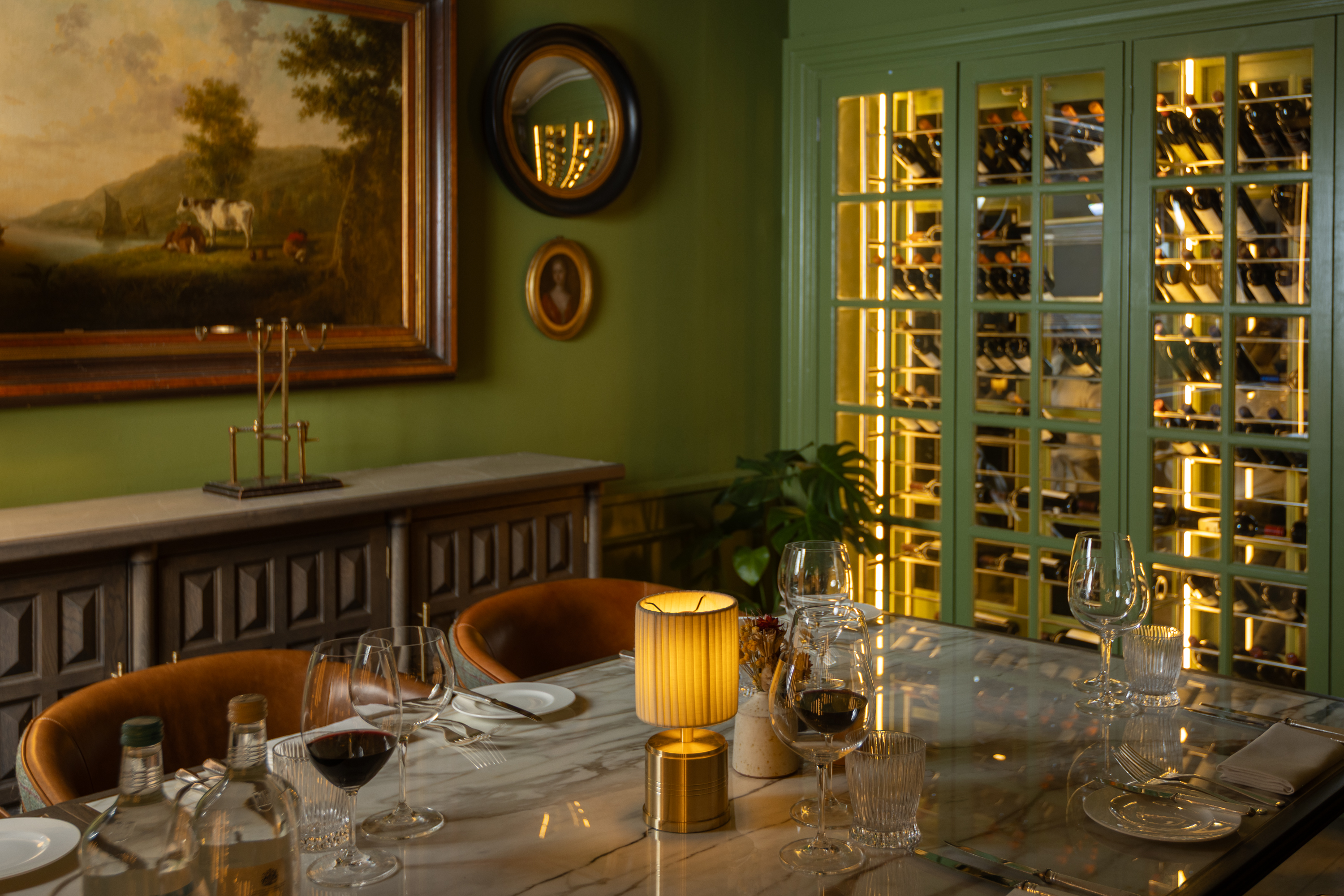 Intimate dining table with wine glasses and a small lamp, set in a green-toned room with illuminated wine cabinets and classic framed artwork