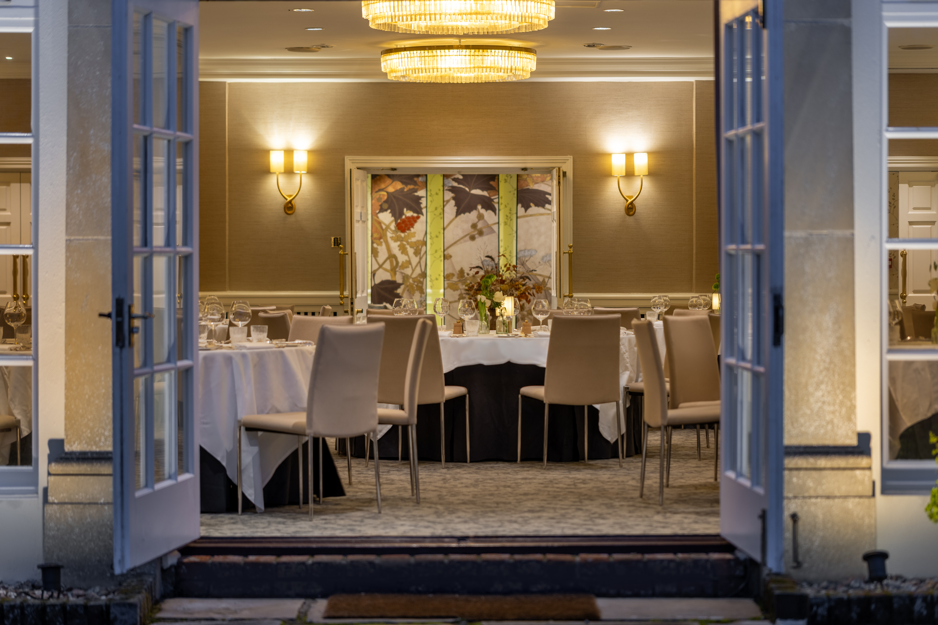 Open French doors revealing an elegant dining room with round tables dressed in white linens, beige chairs, warm wall sconces, and chandelier lighting