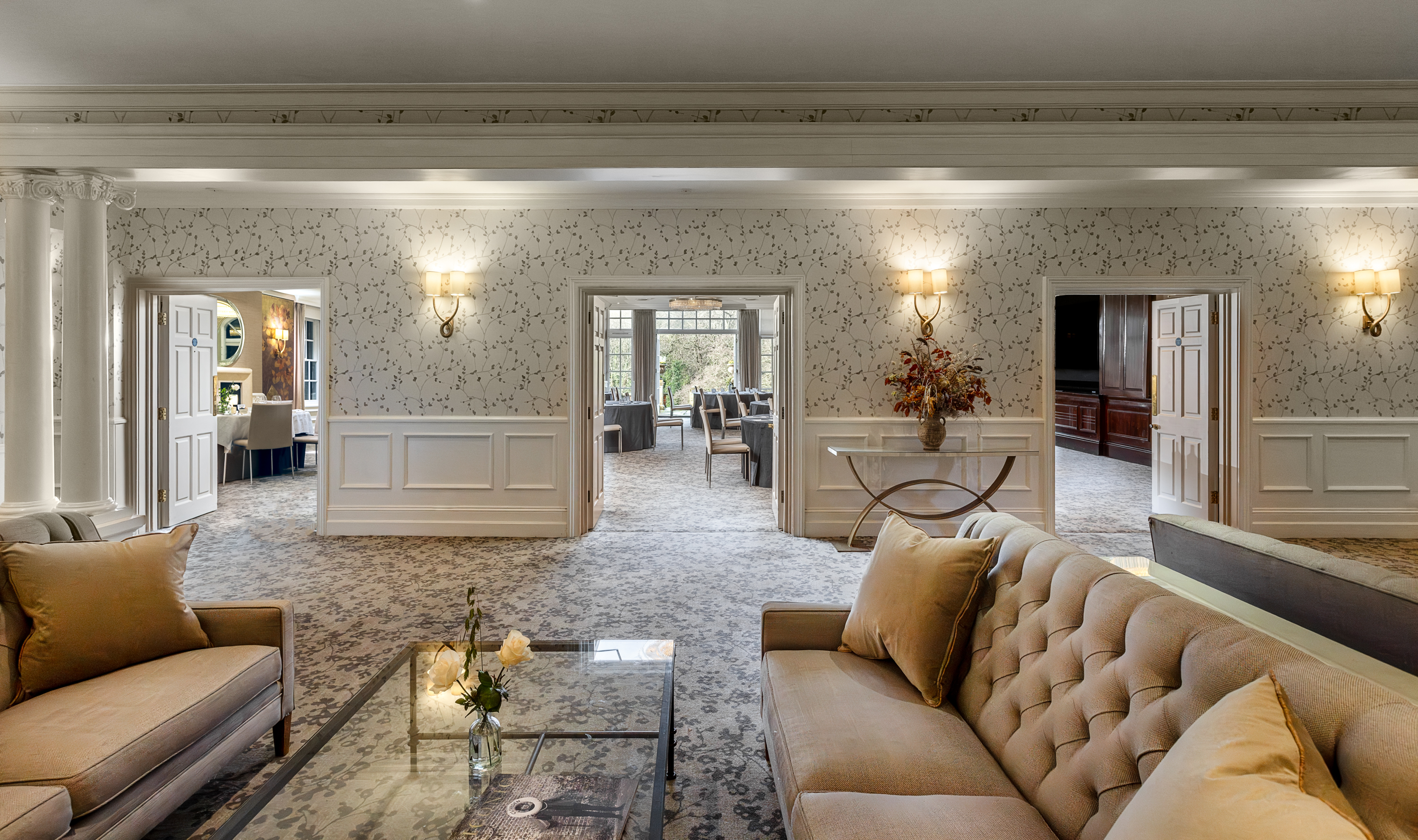 Bright lounge area with patterned walls, soft lighting, and cushioned sofas, opening into an elegant event space with round tables and large windows