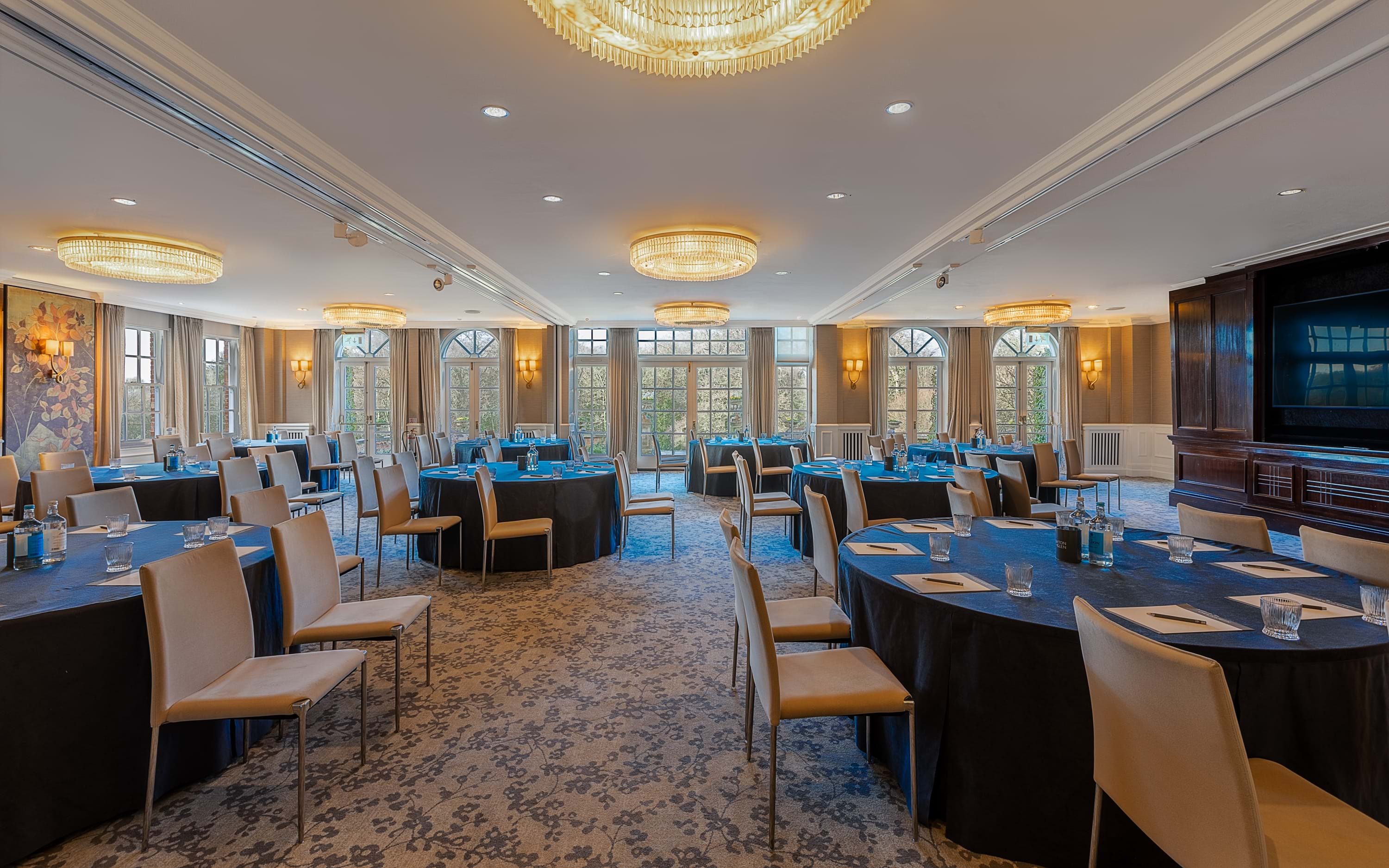 Spacious event room with round tables set for a meeting, beige chairs, large windows, and multiple chandelier lights