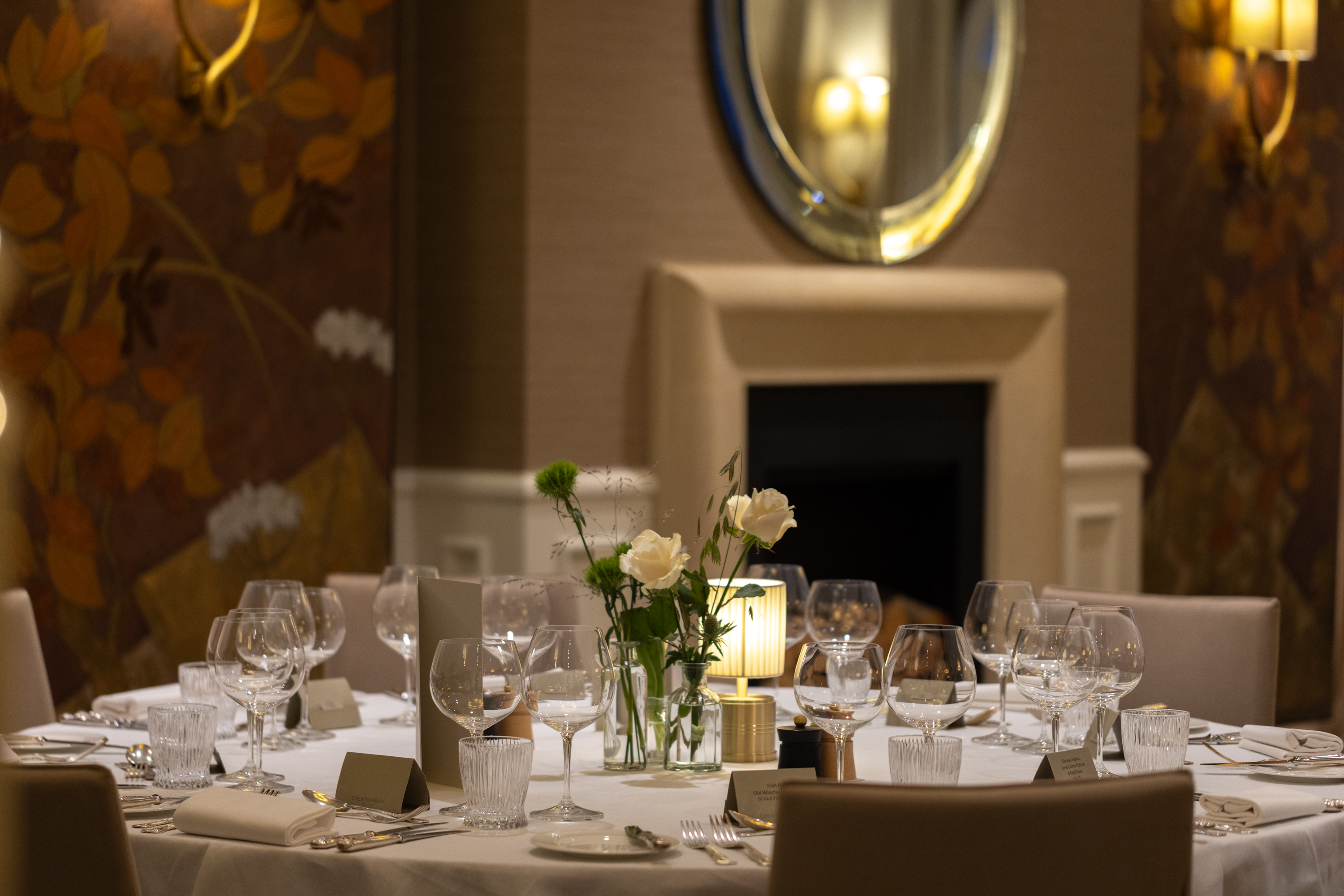 Elegant dinner table set with glassware, cutlery, place cards, and a floral centerpiece, with warm lighting, patterned walls, and a mirror above a fireplace