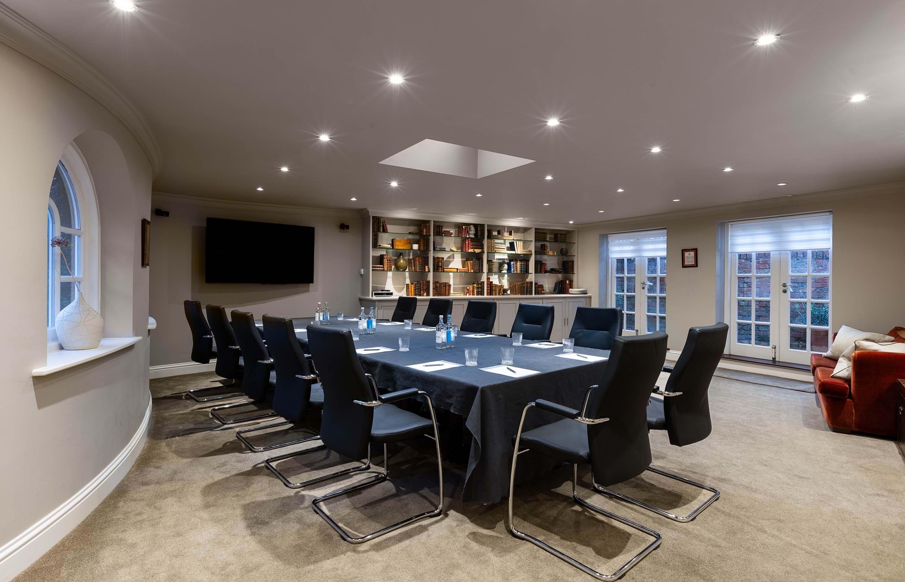 Modern boardroom with a long table set with notepads, glasses, and water bottles, surrounded by black chairs, with bookshelves and large windows along the walls