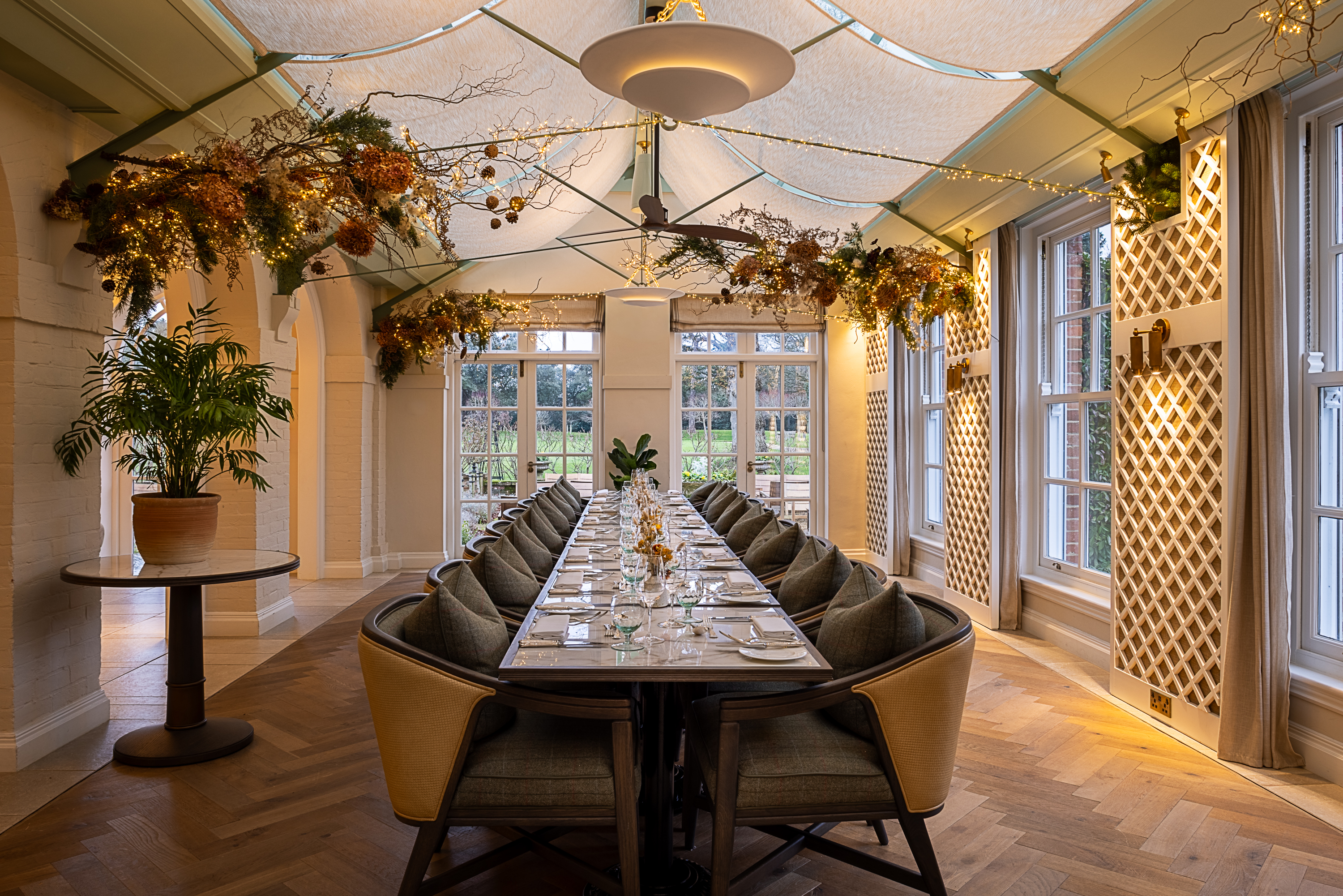 Long private dining table set with glassware and cutlery in a bright conservatory-style room decorated with hanging foliage and large windows overlooking the gardens