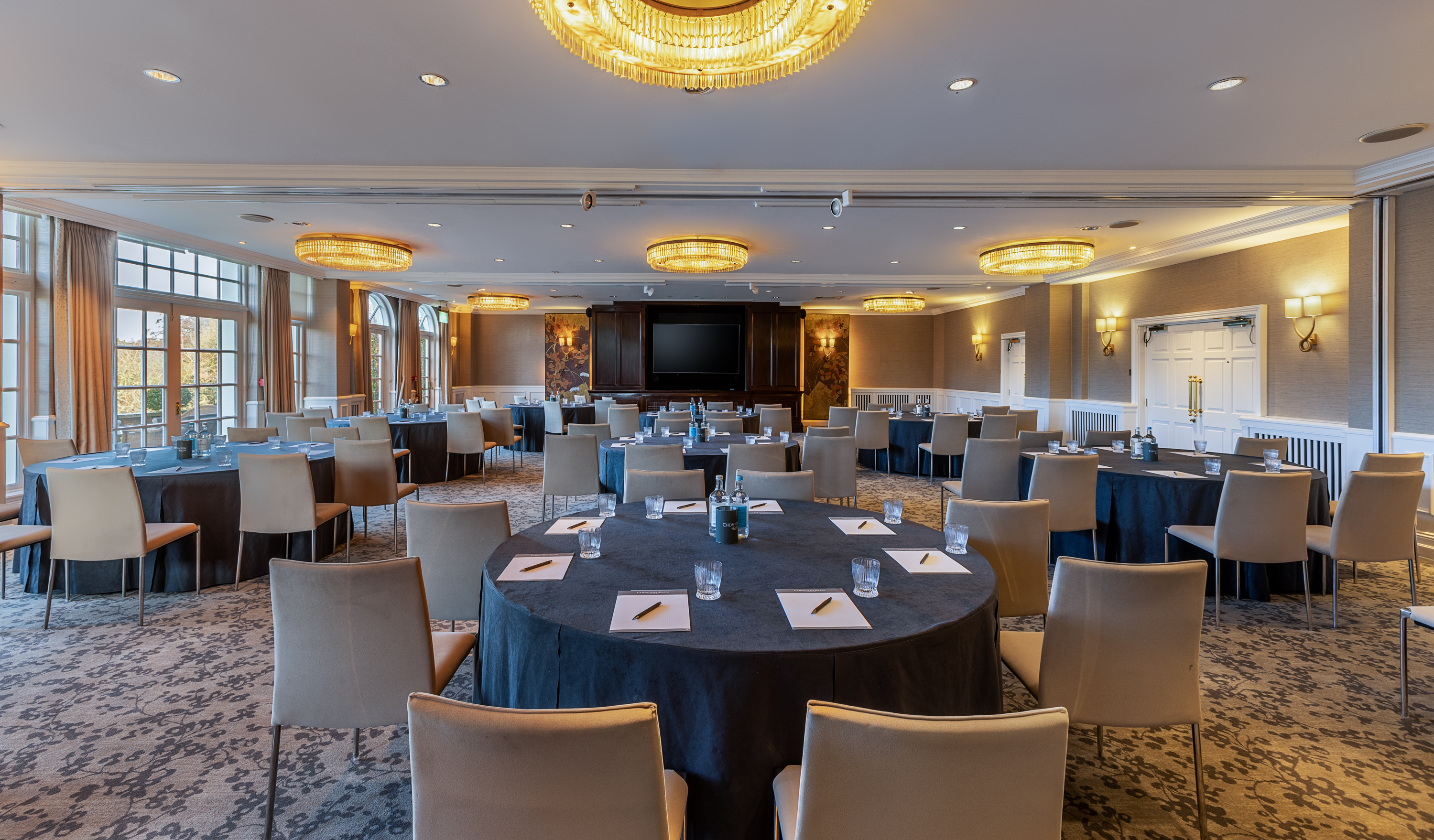 Large conference room with round tables set for a meeting, featuring beige chairs, notepads, water bottles, and warm chandelier lighting